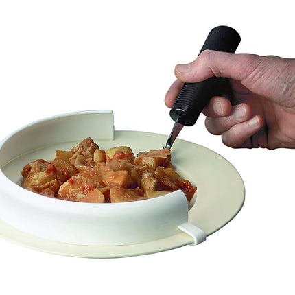 Flexible Food Guard attached to a plate aiding one-handed eating, with a hand holding a utensil, preventing food spillage.