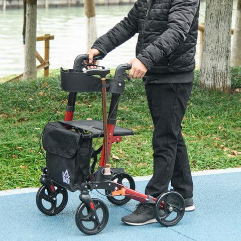 Person using a red 4 wheel rollator walker with a seat and storage bag in a park setting.